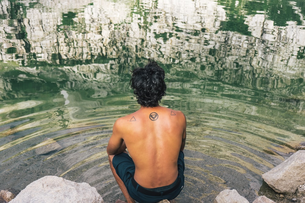 uomo in pantaloncini neri seduto sulla roccia vicino allo specchio d'acqua durante il giorno