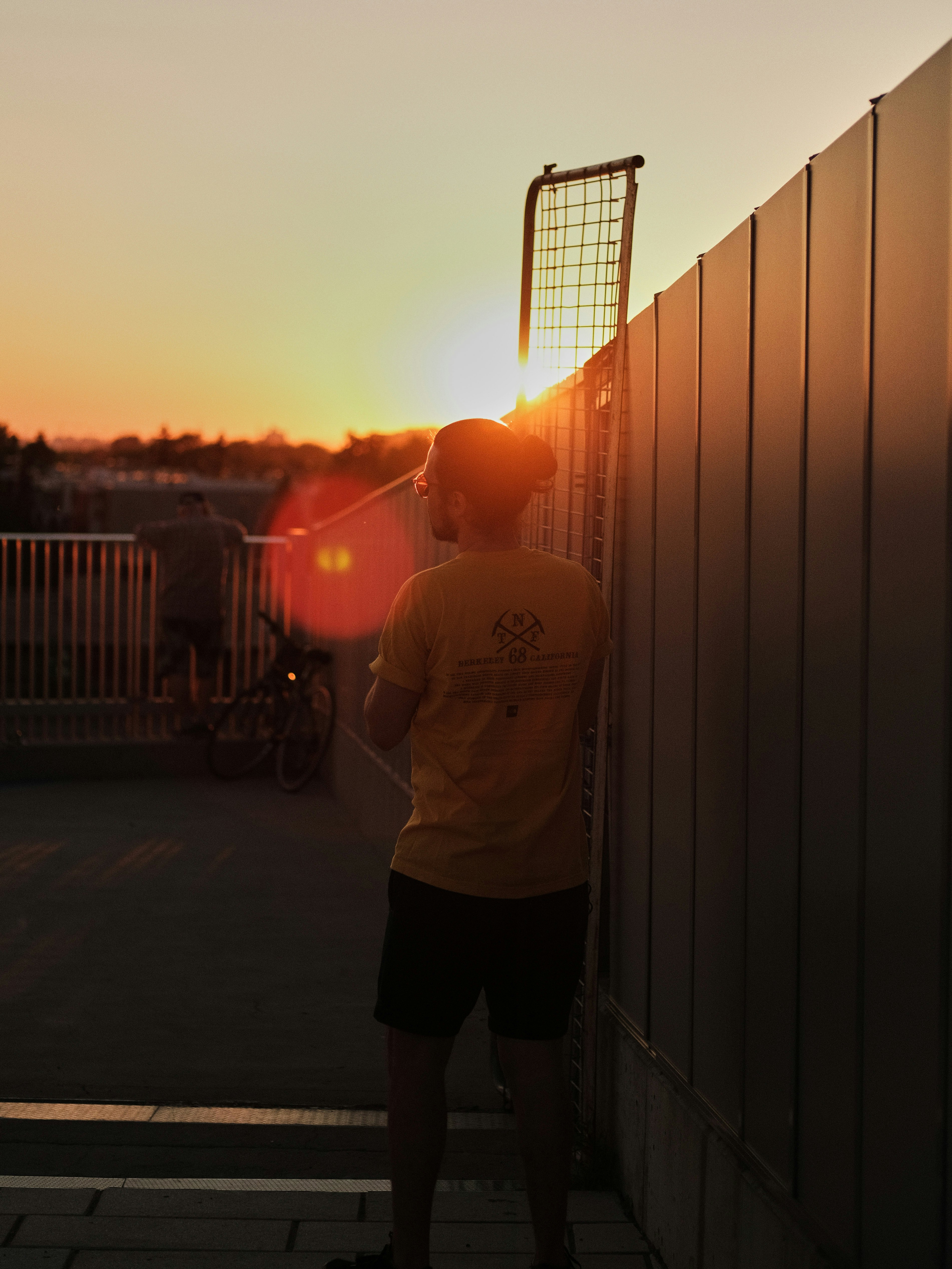 man in white crew neck t-shirt and black pants standing beside fence during sunset
