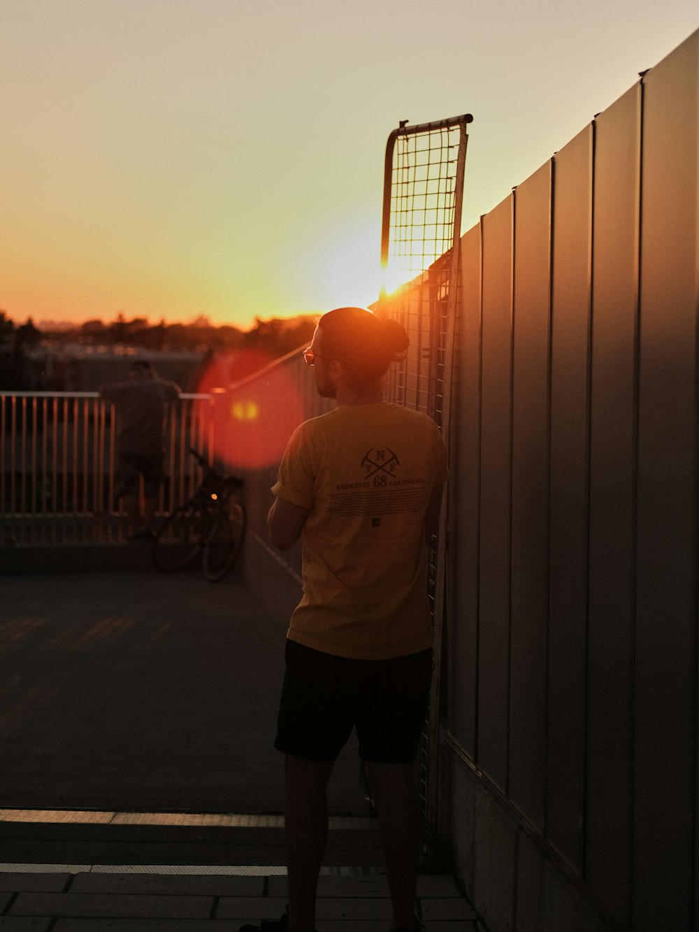 man in white crew neck t-shirt and black pants standing beside fence during sunset