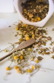 Dried herbs and flowers are scattered across a surface, spilling out of a white container. In the midst of the natural elements, a small wooden spoon with 'made with love' written on it is visible.