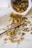 Dried herbs and flowers are scattered across a surface, spilling out of a white container. In the midst of the natural elements, a small wooden spoon with 'made with love' written on it is visible.