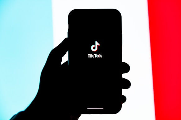 A silhouetted hand holds a smartphone displaying the TikTok logo on its screen, set against a tricolor background of light blue, white, and red.