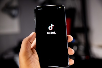 A hand holding a smartphone displaying the TikTok logo against a dark screen, with a blurred background. The hand is positioned prominently in the foreground, and the logo is centered on the phone's display.
