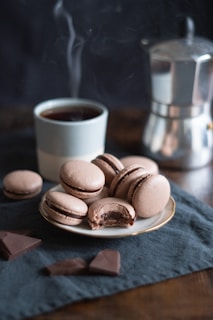 A plate of chocolate macarons with one macaron showing a bite, surrounded by additional pieces of chocolate on a dark cloth. A steaming cup of coffee is placed beside the plate, with a moka pot visible in the background.