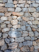 A tightly-packed stone wall composed of irregularly shaped rocks, exhibiting various shades of gray, brown, and tan. The stones are stacked in a manner that suggests stability and robustness, with textures highlighting the rough, natural surface of the material.