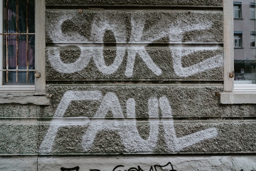 Graffiti with the words 'SOKE FAUL' is spray-painted in white on a textured, gray concrete wall. There are windows with metal bars on the left and a regular window on the right of the wall.