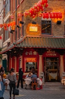 A bustling street corner featuring a Chinese restaurant adorned with red lanterns. People are seen dining at outdoor tables under hanging lanterns with a traditional architectural style. The sign reads 'Lotus Garden' and 'Dim Sum Daily', conveying a lively atmosphere.