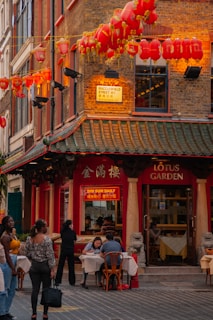 A bustling street corner featuring a Chinese restaurant adorned with red lanterns. People are seen dining at outdoor tables under hanging lanterns with a traditional architectural style. The sign reads 'Lotus Garden' and 'Dim Sum Daily', conveying a lively atmosphere.