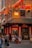A bustling street corner featuring a Chinese restaurant adorned with red lanterns. People are seen dining at outdoor tables under hanging lanterns with a traditional architectural style. The sign reads 'Lotus Garden' and 'Dim Sum Daily', conveying a lively atmosphere.