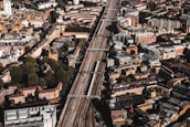 Aerial view of a densely populated urban area with a prominent railway line running through the center. The cityscape features a mix of residential and commercial buildings, mostly characterized by brick and stone facades. Green spaces are interspersed among the structures, providing a contrast to the urban density.