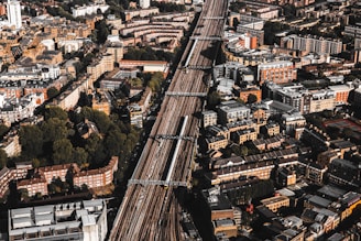 Aerial view of a densely populated urban area with a prominent railway line running through the center. The cityscape features a mix of residential and commercial buildings, mostly characterized by brick and stone facades. Green spaces are interspersed among the structures, providing a contrast to the urban density.