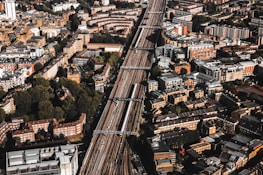 Aerial view of a densely populated urban area with a prominent railway line running through the center. The cityscape features a mix of residential and commercial buildings, mostly characterized by brick and stone facades. Green spaces are interspersed among the structures, providing a contrast to the urban density.