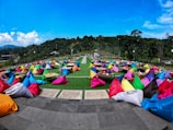 Numerous colorful bean bags placed on a spacious outdoor patio with small tables set on green grass. The scene is surrounded by lush greenery and trees under a bright blue sky. String lights are visible overhead, suggesting a festive or casual gathering atmosphere.
