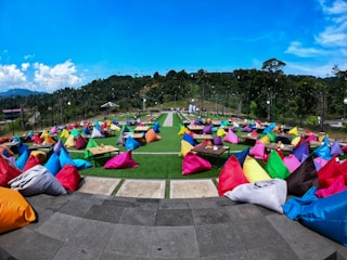 Numerous colorful bean bags placed on a spacious outdoor patio with small tables set on green grass. The scene is surrounded by lush greenery and trees under a bright blue sky. String lights are visible overhead, suggesting a festive or casual gathering atmosphere.