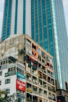 Tall buildings with numerous commercial signs and advertisements. A modern glass skyscraper stands next to an older structure with visible wear and multiple shop signs. The older building hosts various businesses, including cafes and boutiques, as indicated by the vibrant signage. Vegetation is visible on some balconies, providing a contrast to the urban environment.