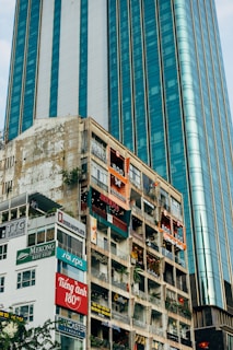 Tall buildings with numerous commercial signs and advertisements. A modern glass skyscraper stands next to an older structure with visible wear and multiple shop signs. The older building hosts various businesses, including cafes and boutiques, as indicated by the vibrant signage. Vegetation is visible on some balconies, providing a contrast to the urban environment.