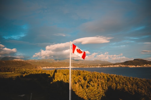 A Canadian flag flies prominently against a backdrop of lush green trees and a large body of water. In the distance, mountains rise under a sky dotted with clouds, casting a serene and picturesque view.