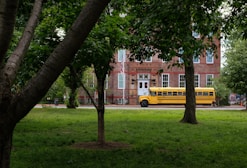 A yellow school bus is parked in front of a historic brick building surrounded by lush green trees and grass. The building has large windows with white frames and a classic architectural style.