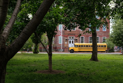 A yellow school bus is parked in front of a historic brick building surrounded by lush green trees and grass. The building has large windows with white frames and a classic architectural style.
