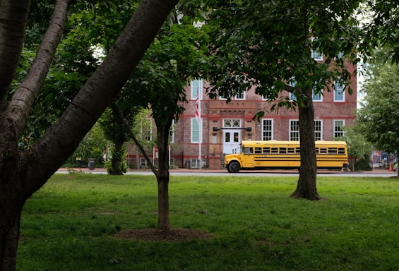 A yellow school bus is parked in front of a historic brick building surrounded by lush green trees and grass. The building has large windows with white frames and a classic architectural style.