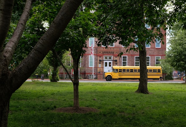 A yellow school bus is parked in front of a historic brick building surrounded by lush green trees and grass. The building has large windows with white frames and a classic architectural style.