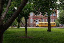 A yellow school bus is parked in front of a historic brick building surrounded by lush green trees and grass. The building has large windows with white frames and a classic architectural style.