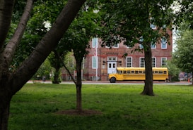 A yellow school bus is parked in front of a historic brick building surrounded by lush green trees and grass. The building has large windows with white frames and a classic architectural style.