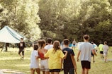 A group of young people are gathered in a grassy area surrounded by lush trees, with several tents visible in the background. Some individuals are standing close together with their arms around each other, creating a sense of camaraderie. Others are walking or standing separately, suggesting an outdoor, possibly camping or team-building activity.