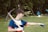 A person wearing a blue sports jersey and gloves is holding a pickaxe. They are standing outdoors on a grassy area with trees in the background, and are pointing towards something. A blue tent is visible in the distance among other camping items.