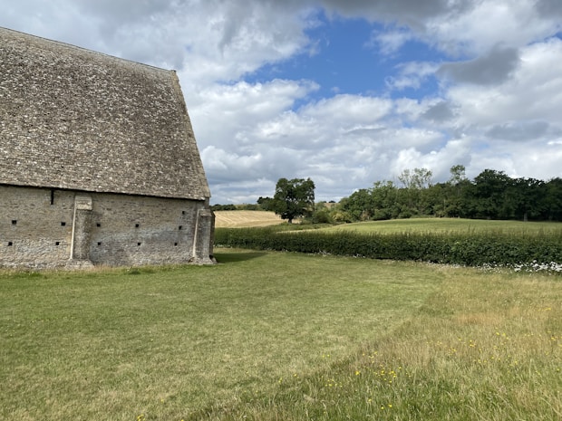 A large, old stone barn with a steeply pitched roof stands next to an expansive field. The scene is surrounded by lush greenery, including hedges, trees, and a neatly mown lawn. The sky above is partially cloudy, allowing patches of blue to be visible.
