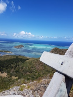 A vast, vibrant seascape with turquoise waters and small islands is visible from a high vantage point. The foreground shows a weathered cross made of stone or concrete. Lush green vegetation covers the slopes below, while white clouds dot the clear blue sky.
