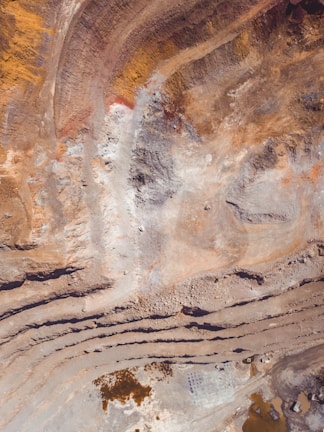 Aerial view of an open-pit mine with visible layers of excavated earth. The terrain is marked by striations and varying earth tones, indicating different mineral deposits. Sparse vegetation or water may be present in certain areas.