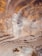 Aerial view of an open-pit mine with visible layers of excavated earth. The terrain is marked by striations and varying earth tones, indicating different mineral deposits. Sparse vegetation or water may be present in certain areas.