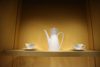 A white porcelain coffee pot with an elegant spout is flanked by two white teacups and saucers. These items are displayed on a beige shelf within a softly lit cabinet, creating a minimalist and clean arrangement.