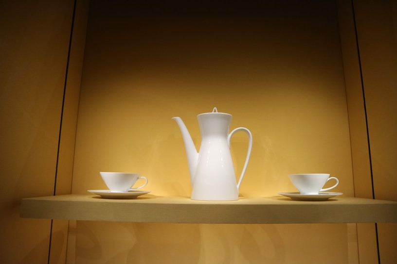A white porcelain coffee pot with an elegant spout is flanked by two white teacups and saucers. These items are displayed on a beige shelf within a softly lit cabinet, creating a minimalist and clean arrangement.