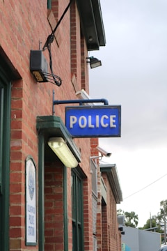 A brick building with a prominent blue sign displaying the word 'POLICE'. Below the sign, there is a plaque indicating it is a police station. The structure features multiple windows and outdoor lighting fixtures. The surrounding environment includes some greenery and additional buildings in the background.