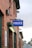A brick building with a prominent blue sign displaying the word 'POLICE'. Below the sign, there is a plaque indicating it is a police station. The structure features multiple windows and outdoor lighting fixtures. The surrounding environment includes some greenery and additional buildings in the background.
