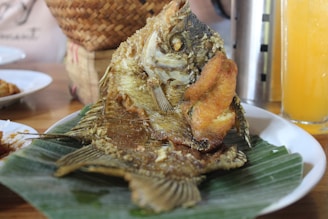A whole fried fish is served on a large green banana leaf placed on a white plate. The fish appears crispy and golden brown with visible fins and tail. Nearby, a glass of orange juice is partially visible along with a rice basket in the background.