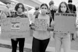 Three people wearing masks hold protest signs at a public demonstration. The signs convey messages supporting social justice movements, including Black Lives Matter and advocating against systematic oppression.