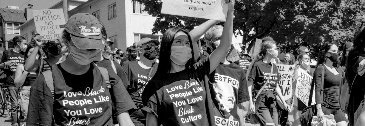 A group of people are participating in a protest or march. They are wearing masks and holding signs with messages advocating for racial justice and equality. Many participants are wearing t-shirts with supportive slogans for Black culture. The setting appears to be an urban area with buildings and trees visible in the background.