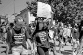 A group of people are participating in a protest or march. They are wearing masks and holding signs with messages advocating for racial justice and equality. Many participants are wearing t-shirts with supportive slogans for Black culture. The setting appears to be an urban area with buildings and trees visible in the background.