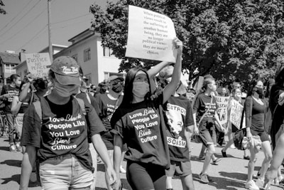 A group of people are participating in a protest or march. They are wearing masks and holding signs with messages advocating for racial justice and equality. Many participants are wearing t-shirts with supportive slogans for Black culture. The setting appears to be an urban area with buildings and trees visible in the background.