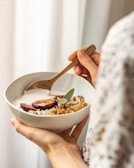 A person holds a bowl filled with yogurt topped with granola, sliced plums, and a sprig of sage. The bowl is in one hand while the other uses a wooden spoon. The setting appears warm and natural with soft lighting.
