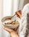 Close-up of a smiling woman holding a bowl of cooked natural grains in a softly lit kitchen.