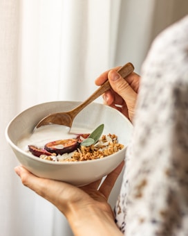 A person holds a bowl filled with yogurt topped with granola, sliced plums, and a sprig of sage. The bowl is in one hand while the other uses a wooden spoon. The setting appears warm and natural with soft lighting.