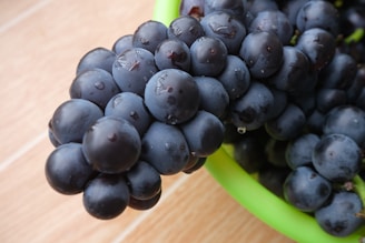 A close-up view of a bunch of dark purple grapes sitting in a green bowl on a light wooden surface. The grapes appear fresh and juicy, with small droplets of water visible on their smooth surfaces.
