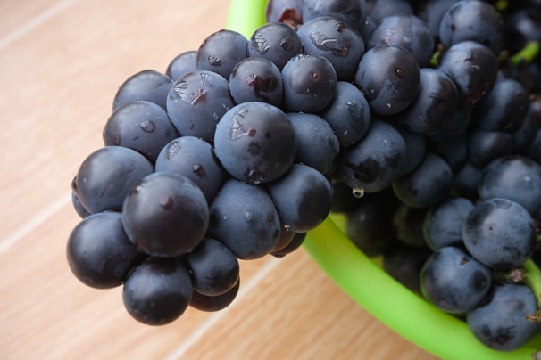 A close-up view of a bunch of dark purple grapes sitting in a green bowl on a light wooden surface. The grapes appear fresh and juicy, with small droplets of water visible on their smooth surfaces.