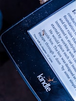 A Kindle e-reader displays text with a line indicating the time left in the chapter. Several small ants are visible on the screen and edges of the Kindle. The lighting gives the image a contrasting effect, highlighting the presence of the ants on the device.