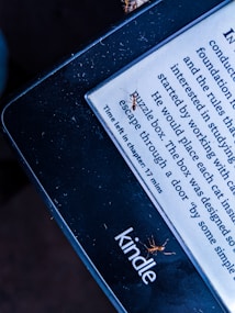 A Kindle e-reader displays text with a line indicating the time left in the chapter. Several small ants are visible on the screen and edges of the Kindle. The lighting gives the image a contrasting effect, highlighting the presence of the ants on the device.