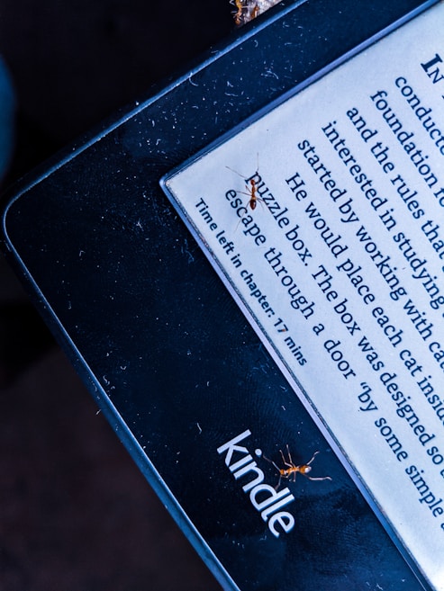 A Kindle e-reader displays text with a line indicating the time left in the chapter. Several small ants are visible on the screen and edges of the Kindle. The lighting gives the image a contrasting effect, highlighting the presence of the ants on the device.